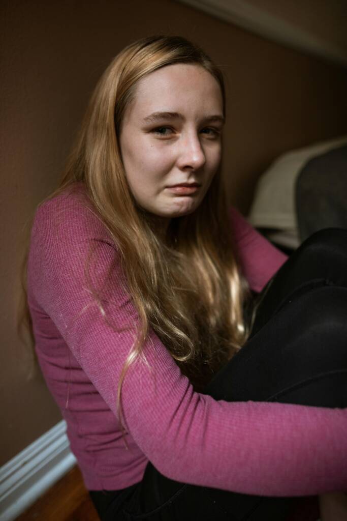 A teenage girl with a distressed expression, wearing a pink sweater, sits indoors.